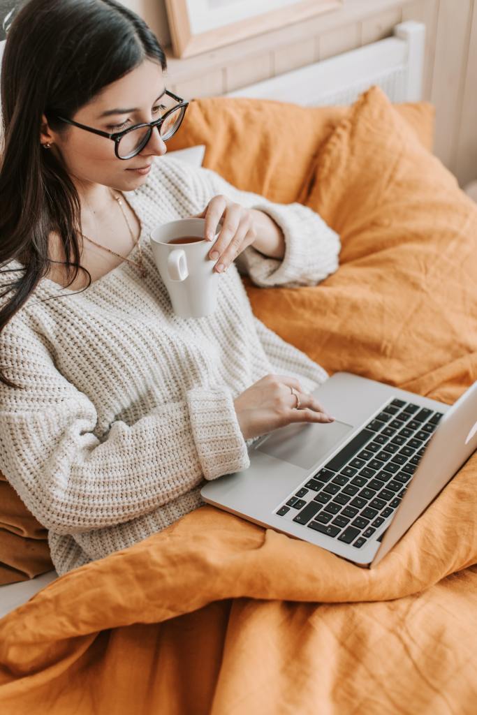 a person with long hair and glasses holds a cup of tea whilst snuggled in bed and looking at a computer. Image (c) Vlada Karpovich.